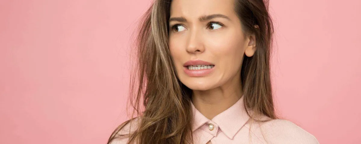 A woman against a pink background biting her lip and glancing sideways with an anxious expression.