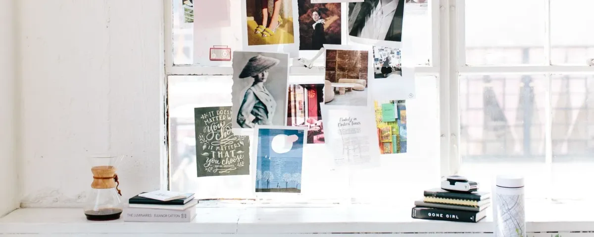 A bright window ledge with a mood board of photographs and prints pinned above it, beside books and a coffee maker.