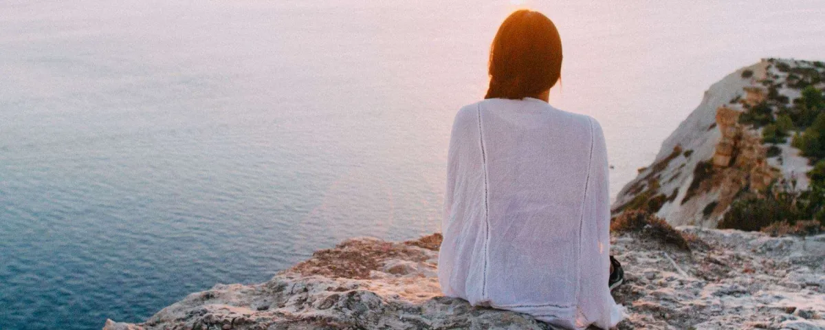 woman sitting on a cliff overlooking the water at sunrise or sunset with the sky and water in soft pastel colors