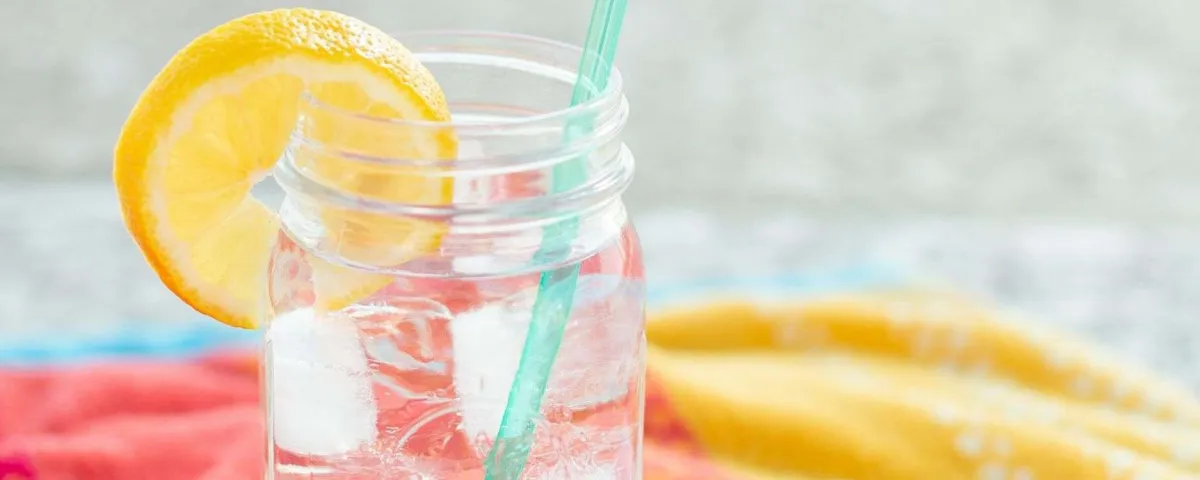 A mason jar of iced water with a lemon slice and a straw, resting on a bright towel by the water.
