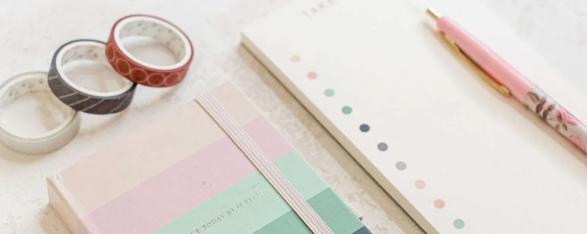 A notebook, a dotted planner page, a pink pen and rolls of washi tape arranged on a pale desk.