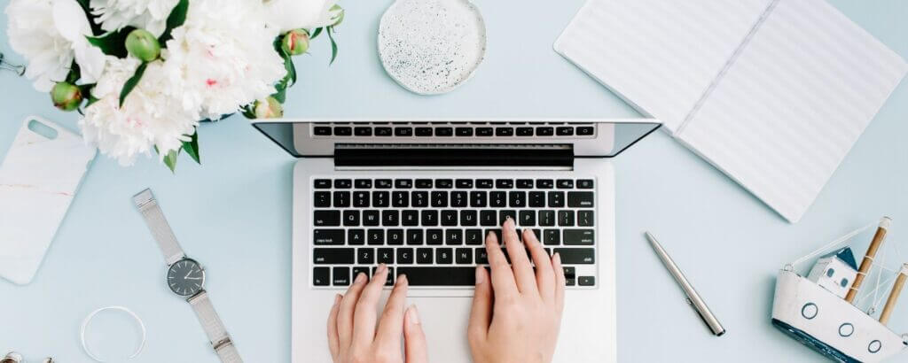 business owner typing on a laptop on a light blue desk with flowers, a watch, and office accessories