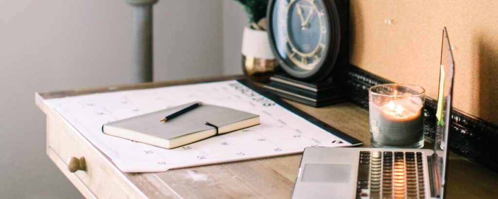 Photo of a desk on an angle showing a computer in the foreground and a notebook, calendar, and clock in the background