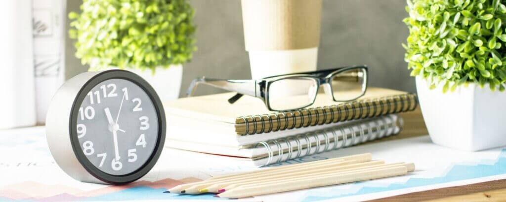 pile of notebooks with glasses on top of them next to a clock with a couple of plants in the background