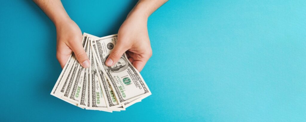 woman's hands holding a bunch of money fanned out like playing cards against a turquoise background
