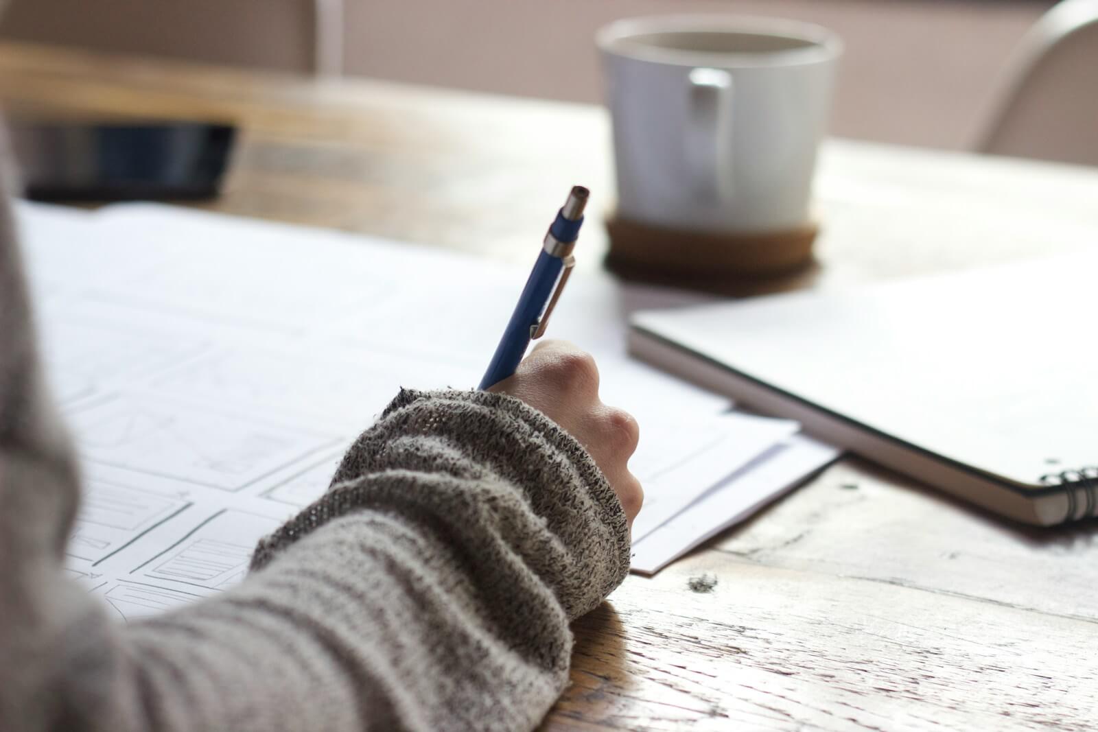 person filling out an assessment on a wooden table near white ceramic mug