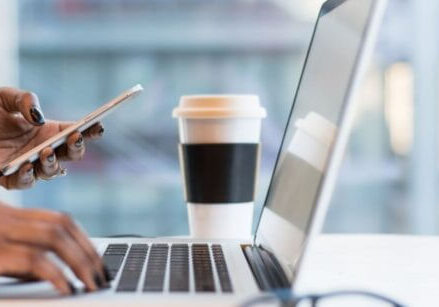 woman's hands holding a phone clicking a link while the other hand is typing on a laptop