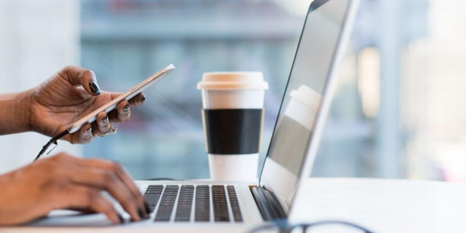 woman's hands holding a phone clicking a link while the other hand is typing on a laptop