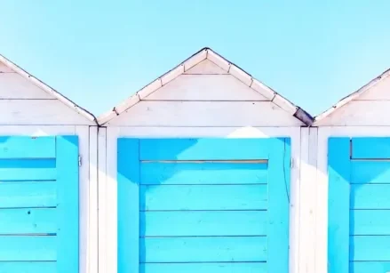 three beach changing rooms with white siding and blue doors. behind them is a blue sky
