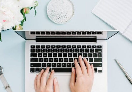 business owner typing on a laptop on a light blue desk with flowers, a watch, and office accessories