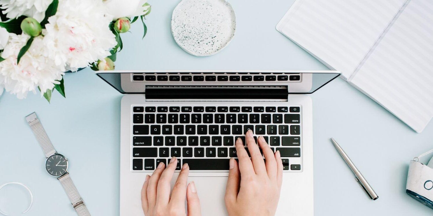 business owner typing on a laptop on a light blue desk with flowers, a watch, and office accessories