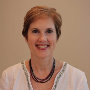 Carole Maier headshot - beautiful woman with short auburn hair wearing a white top and a beaded necklace.