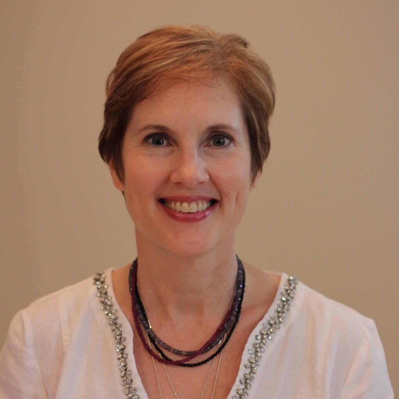 Carole Maier headshot - beautiful woman with short auburn hair wearing a white top and a beaded necklace.