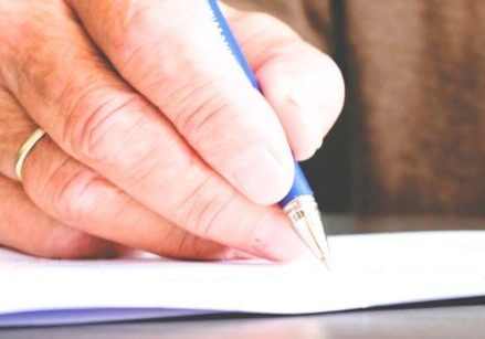 old man's had with a wedding ring holding a pen and writing in a journal