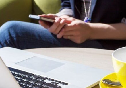 woman holding a cell phone while sitting at her laptop next to a yellow coffee mug