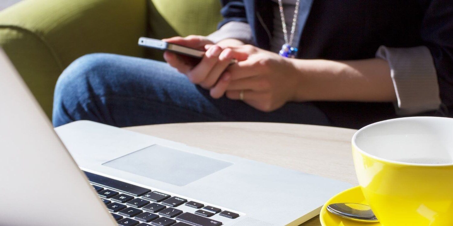woman holding a cell phone while sitting at her laptop next to a yellow coffee mug