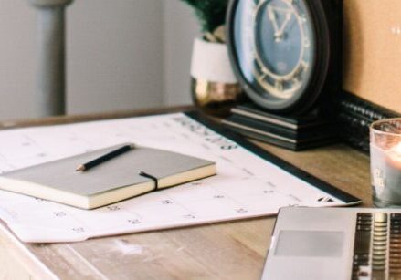 Photo of a desk on an angle showing a computer in the foreground and a notebook, calendar, and clock in the background