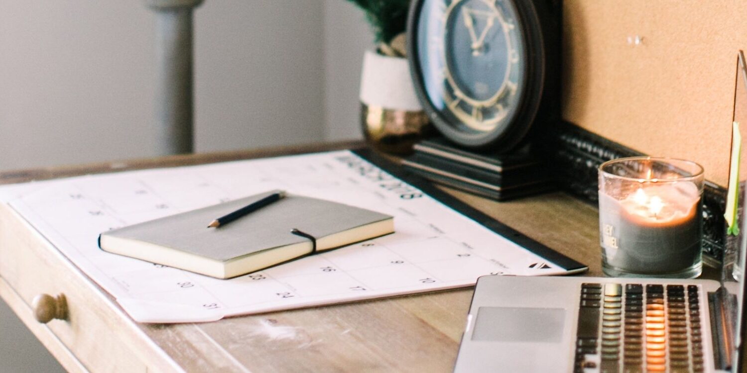 Photo of a desk on an angle showing a computer in the foreground and a notebook, calendar, and clock in the background