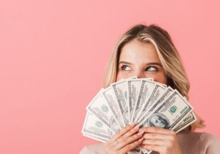 woman peeking with smiling eyes over the top of a handful of money fanned out over a pink background