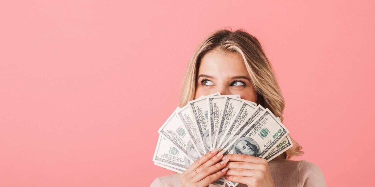 woman peeking with smiling eyes over the top of a handful of money fanned out over a pink background