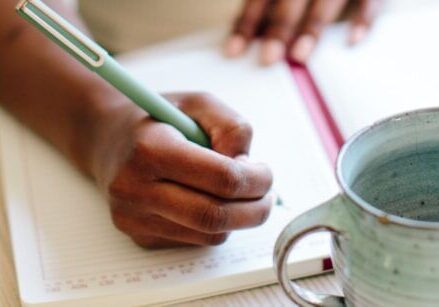 African American woman's hand holding a pen writing in a journal with a ceramic mug on the table