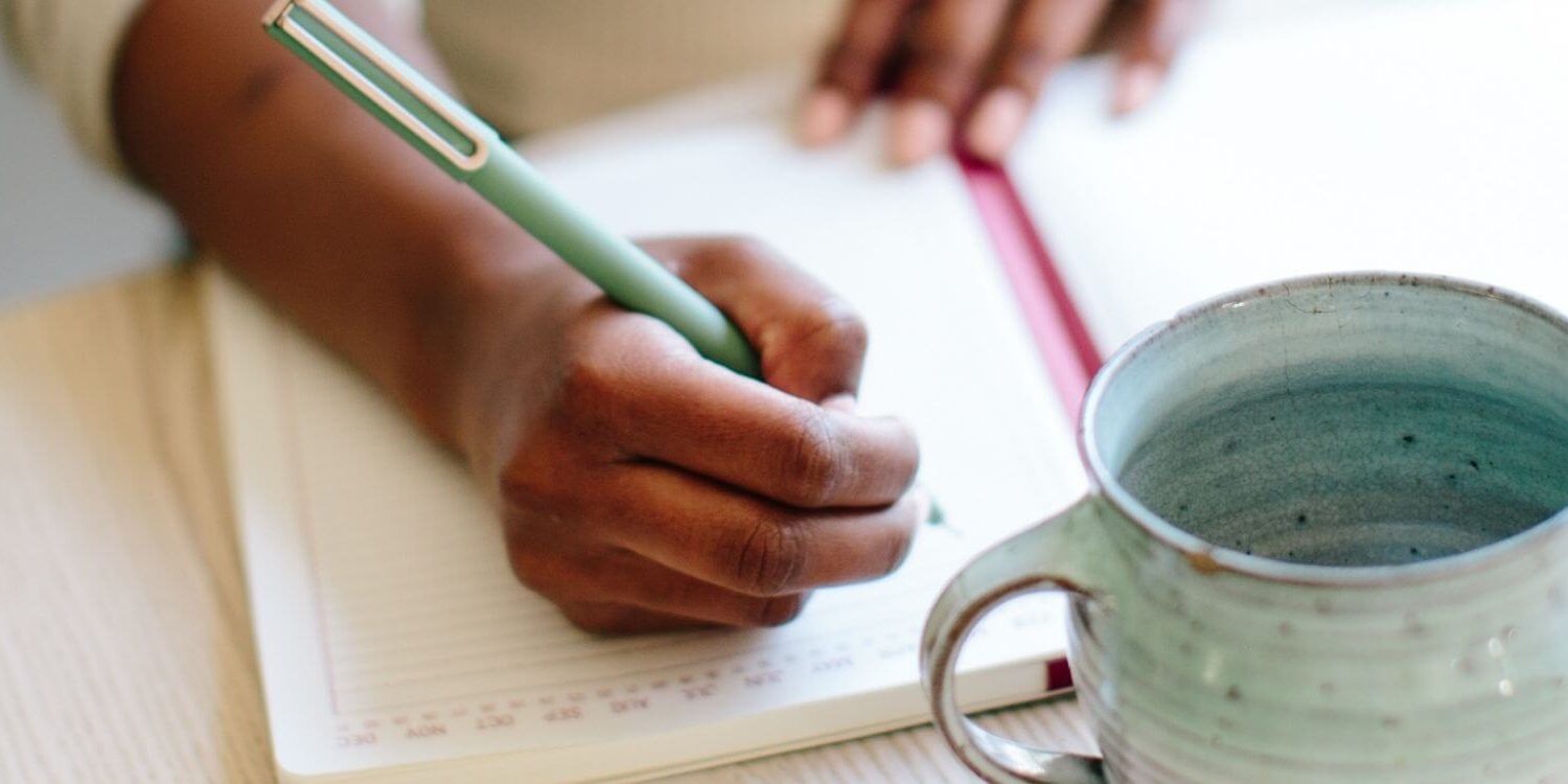 African American woman's hand holding a pen writing in a journal with a ceramic mug on the table