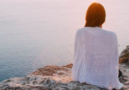 woman sitting on a cliff overlooking the water at sunrise or sunset with the sky and water in soft pastel colors