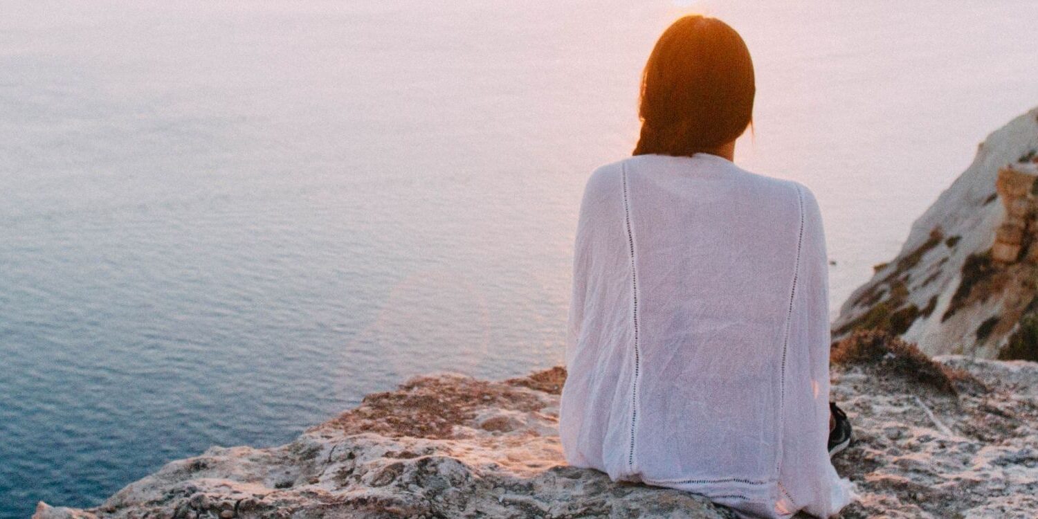 woman sitting on a cliff overlooking the water at sunrise or sunset with the sky and water in soft pastel colors