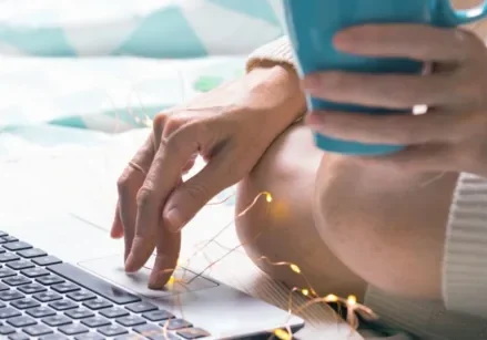 woman sitting on the floor typing on a laptop with one hand and holding a turquoise coffee mug with the other hand