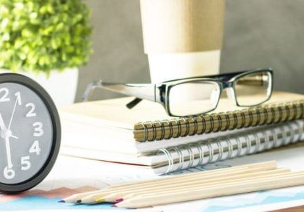 pile of notebooks with glasses on top of them next to a clock with a couple of plants in the background