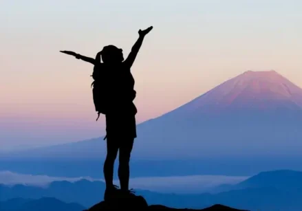 Woman wearing a backpack looking out at purple mountains with her arms up showing success