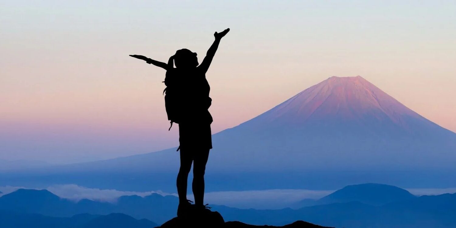 Woman wearing a backpack looking out at purple mountains with her arms up showing success