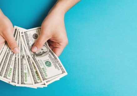 woman's hands holding a bunch of money fanned out like playing cards against a turquoise background