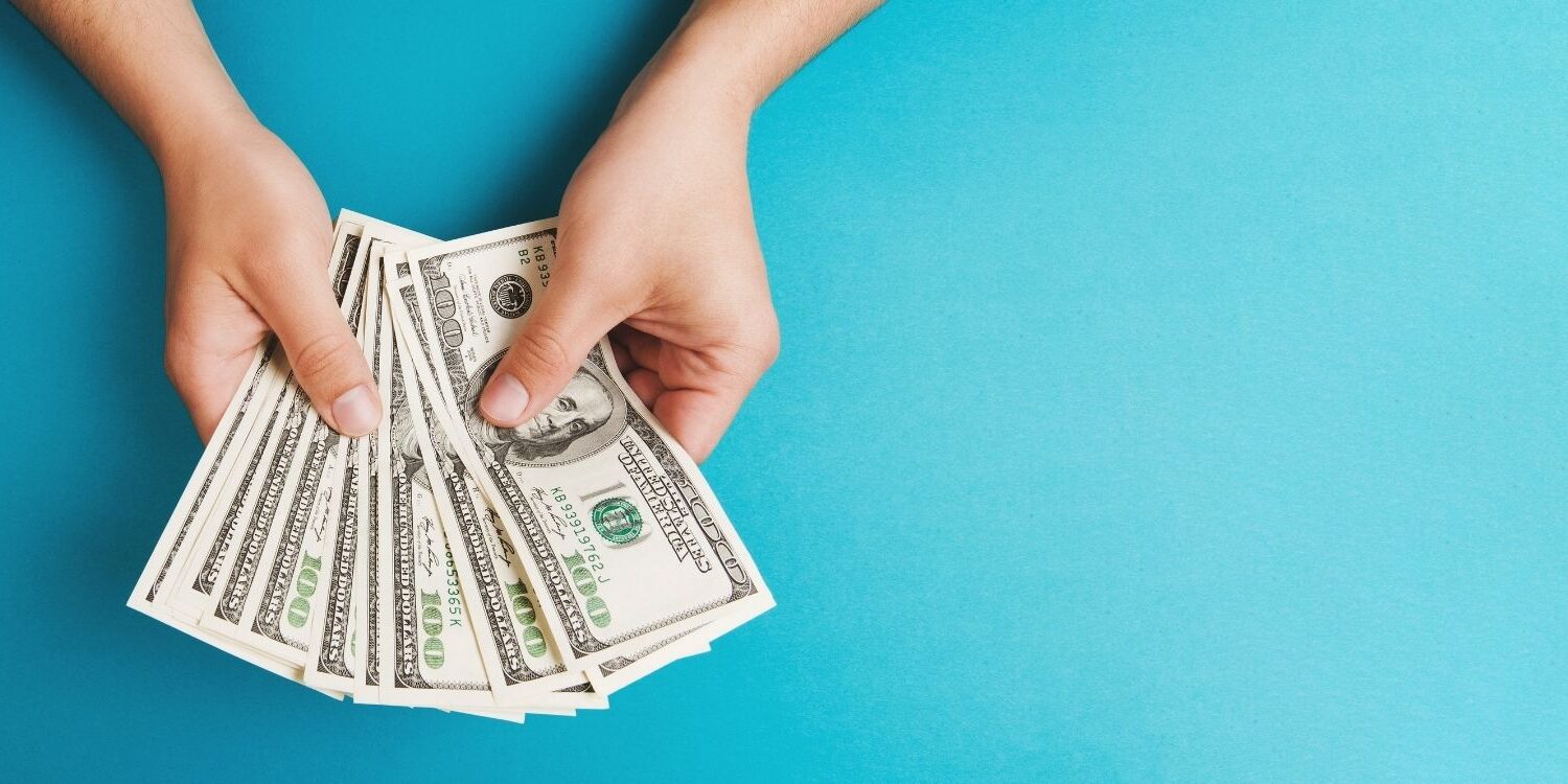 woman's hands holding a bunch of money fanned out like playing cards against a turquoise background