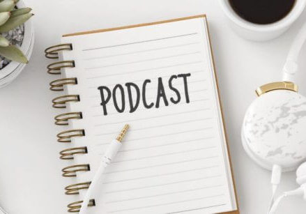 notebook on a desk with the word podcast written at the top of the page. There are headphones, a coffee mug, and plant on the desk.