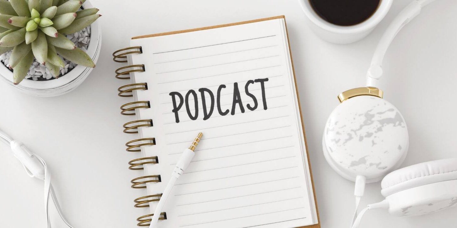 notebook on a desk with the word podcast written at the top of the page. There are headphones, a coffee mug, and plant on the desk.
