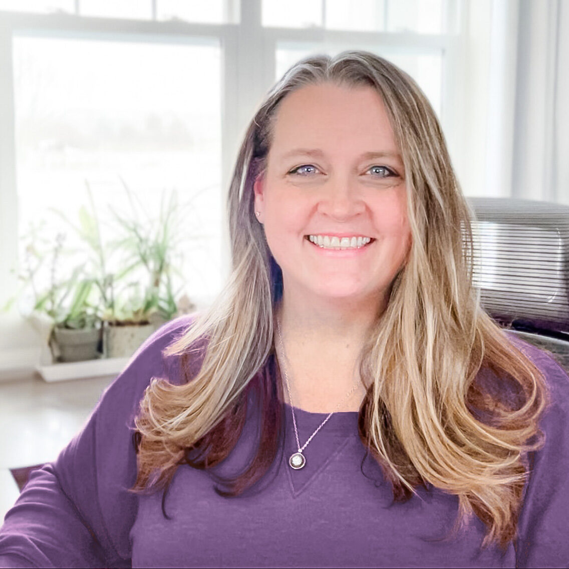 heather stephens: woman with long brown hair wearing a purple sweatshirt sitting at her desk.